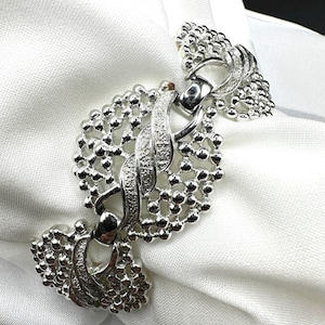 May include: A silver-toned napkin ring with an intricate design. The ring features a central oval shape with a textured, interwoven pattern and is flanked by two decorative, beaded sections. The ring is set against a white fabric background.