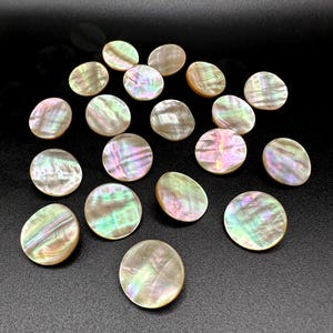 Könnte beinhalten: Eine Sammlung schillernder, runder Perlmuttknöpfe, die auf einer schwarzen Oberfläche verstreut sind. Die Knöpfe zeigen eine Reihe von Farben, darunter Rosa, Grün und Gold, mit einem perlmuttartigen Glanz. Die Knöpfe variieren leicht in Größe und Form.