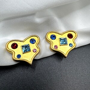 May include: A pair of gold-coloured earrings with a heart-shaped design. Each earring has a central square blue gem, surrounded by red, blue, and pink gems. The earrings are displayed on white fabric.