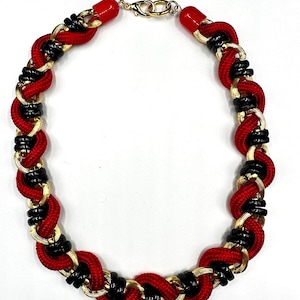 Könnte beinhalten: Eine Halskette in Rot, Schwarz und Gold. Sie hat ein geflochtenes Design mit rotem Seil, schwarzen Akzenten und goldfarbenen Metallgliedern. Die Halskette hat einen goldfarbenen Verschluss und rote Endkappen.