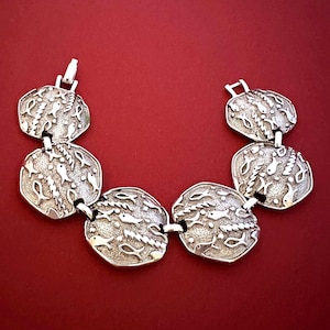 May include: A silver-toned bracelet with six circular links. Each link features a detailed design of fish and aquatic plants. The bracelet is displayed against a red background, highlighting its intricate details and metallic sheen.
