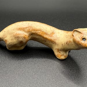 Op de afbeelding: Een keramiek beeldje van een wezel, geschilderd in tinten bruin en beige. De wezel is geposeerd in een rennende houding met zijn hoofd naar de zijkant gedraaid.