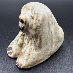 Puede incluir: Una pequeña escultura de cerámica de un perro sentado con las patas cruzadas. El perro es blanco con marcas marrones y tiene la nariz y la lengua negras.