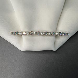Può includere: Una spilla a barra d'argento con strass iridescenti e perle bianche. La spilla è su uno sfondo di tessuto bianco.
