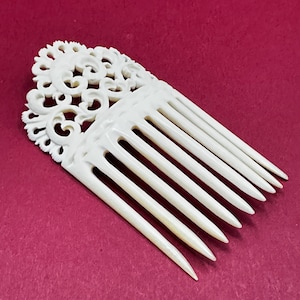 May include: Ivory-colored hair comb with intricate floral and scrollwork design. The comb features a decorative top section and long, evenly spaced teeth. The background is a solid maroon color.