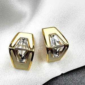 Puede incluir: Un par de pendientes dorados con un diseño geométrico. Cada pendiente presenta un cristal transparente y facetado engastado en un marco de esmalte color crema, contorneado con oro. Los pendientes están sobre un fondo de tela blanca.
