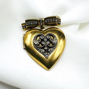 Peut inclure: Broche en forme de cœur de style vintage, en métal doré. Le fermoir est orné d'un nœud décoratif avec des accents de cristaux transparents et d'un motif de filigrane détaillé. Le médaillon s'ouvre pour révéler un espace pour une petite photo ou un souvenir.