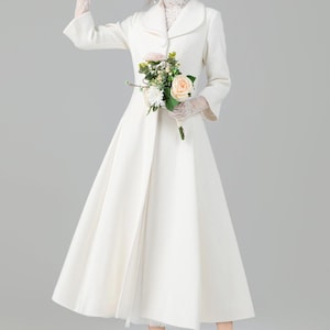 May include: A white coat with a flared skirt, worn with lace gloves and a bouquet of flowers. The coat has a classic design with a rounded collar and long sleeves. The model is wearing white heels.