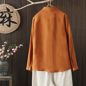May include: A long-sleeved, rust-colored linen shirt with a button-down collar. The shirt is hanging on a wooden hanger against a wood-paneled wall.