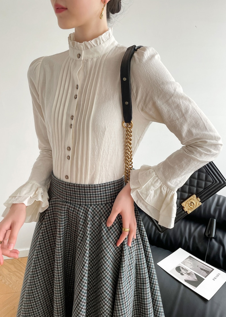 May include: A white long-sleeved blouse with a high ruffled collar, button-up front, and pleated detail. The blouse has ruffled cuffs and is paired with a black and white plaid skirt.