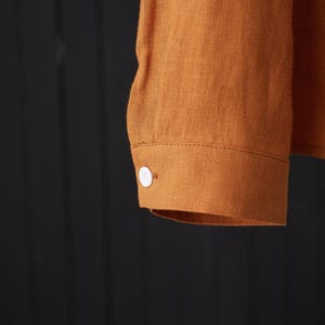 May include: A long-sleeved shirt with a button cuff in a rust orange color. The shirt is made of a linen fabric.