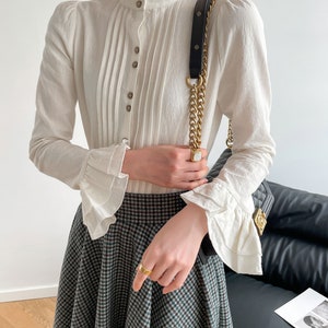 May include: A white long-sleeved blouse with a high ruffled collar and button-up front. The blouse has pleated detailing and ruffled cuffs. The model is wearing a black and white plaid skirt.