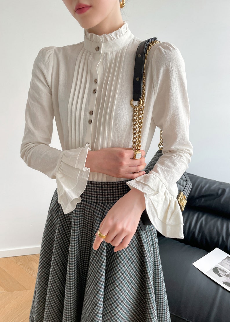 May include: A white long-sleeved blouse with a high ruffled collar and button-up front. The blouse has pleated detailing and ruffled cuffs. The model is wearing a black and white plaid skirt.