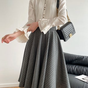 May include: A white blouse with a ruffled collar and long sleeves, paired with a black and green plaid skirt. The skirt is a-line and falls to the knees. The woman is wearing a black quilted handbag with a gold chain strap.
