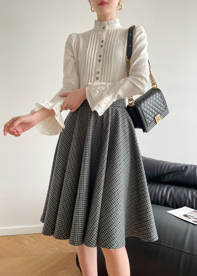 May include: A white blouse with a ruffled collar and long sleeves, paired with a black and green plaid skirt. The skirt is a-line and falls to the knees. The woman is wearing a black quilted handbag with a gold chain strap.