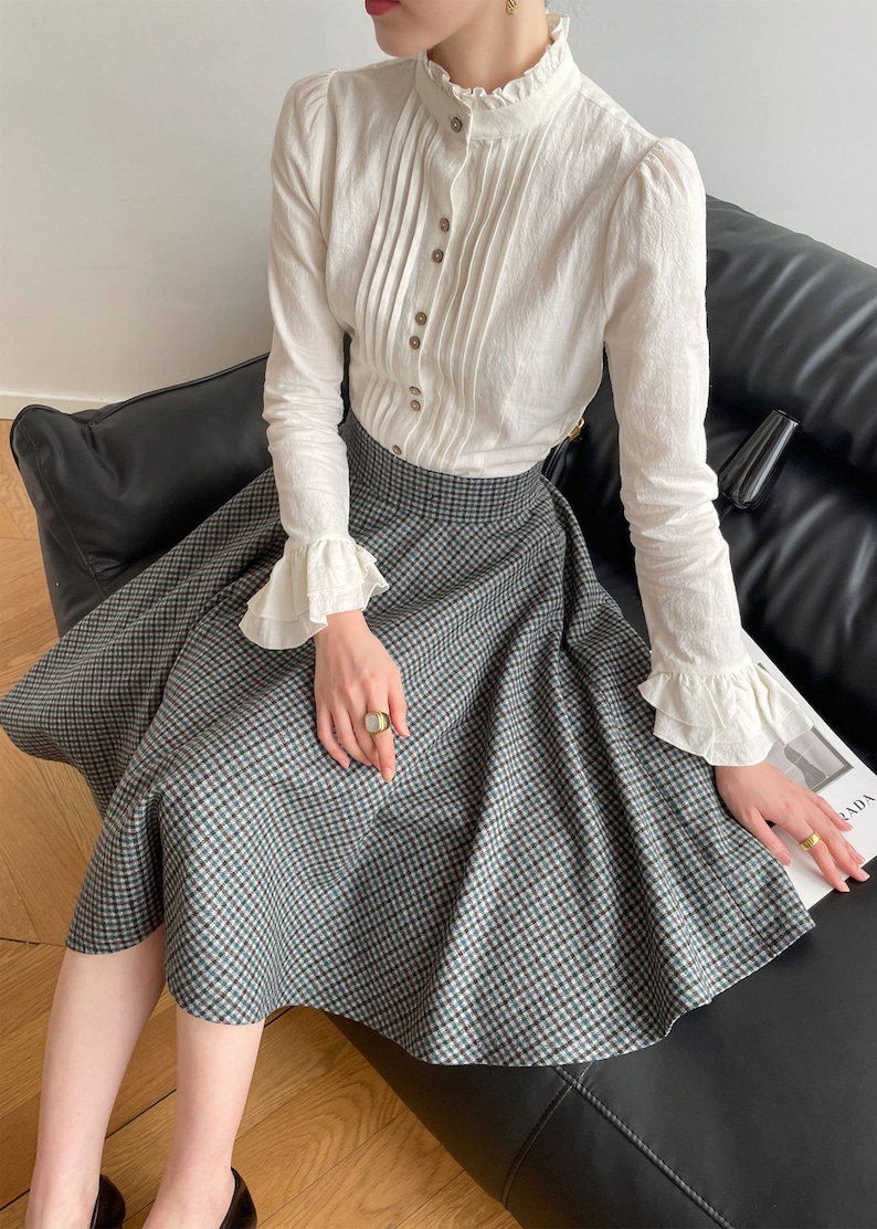 May include: A white long-sleeved blouse with a ruffled collar and cuffs, paired with a green and black plaid skirt. The blouse has a button-up front and a pleated design.