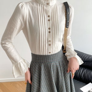 May include: A white long-sleeved blouse with a ruffled collar and button-up front. The blouse has a pleated front and ruffled cuffs. The model is wearing a plaid skirt.