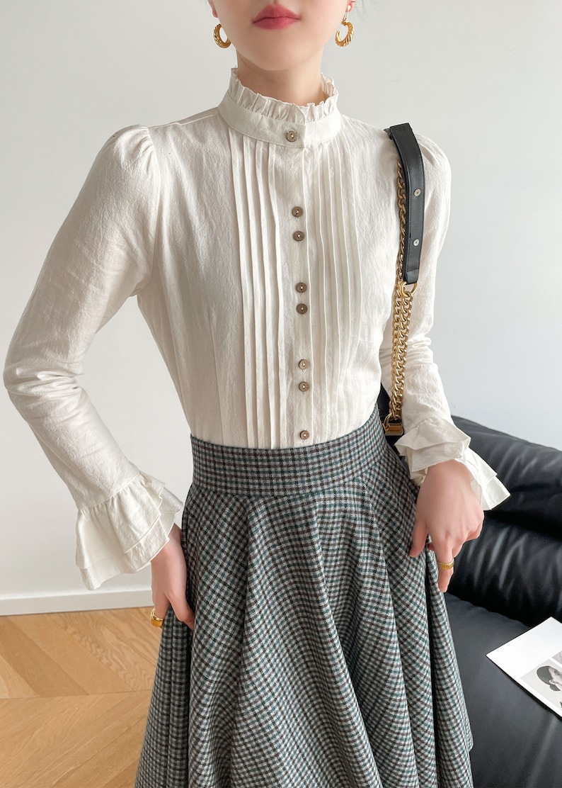 May include: A white long-sleeved blouse with a ruffled collar and button-up front. The blouse has a pleated front and ruffled cuffs. The model is wearing a plaid skirt.