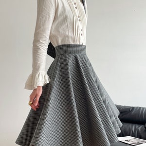 May include: A white long-sleeved blouse with a ruffled collar and a high neckline. The blouse has a button-up front and is paired with a black and white checkered skirt.