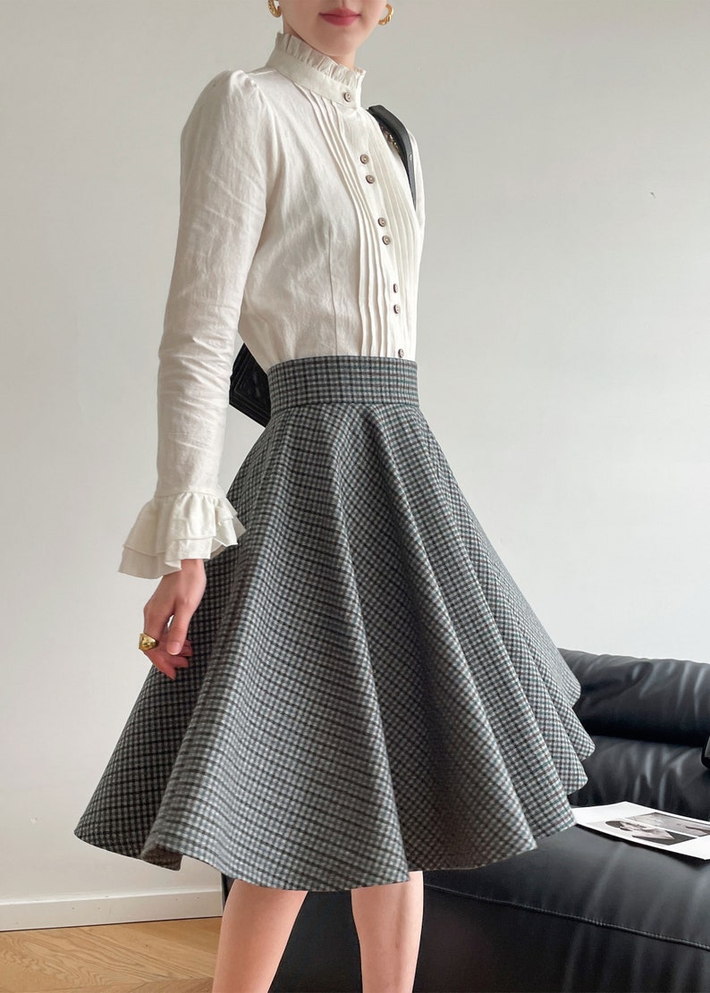 May include: A white long-sleeved blouse with a ruffled collar and a high neckline. The blouse has a button-up front and is paired with a black and white checkered skirt.