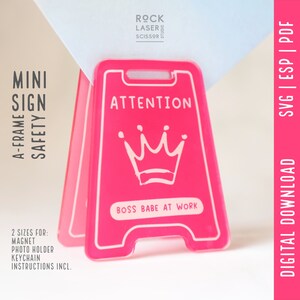 Puede incluir: Un mini letrero rosa en forma de A con texto blanco. El letrero dice "ATTENTION" y "BOSS BABE AT WORK", con un gráfico de corona. Está diseñado para un imán, un portafotos y un llavero.
