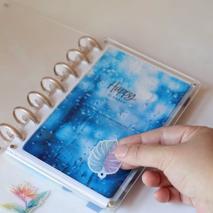 May include: A clear Happy Planner with a blue and white cover, featuring the text "Happy Planner". A hand is placing a leaf-shaped sticker on the planner. Additional floral stickers are also visible.