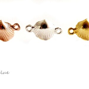 May include: Three seashell-shaped jewelry connectors in rose gold, silver, and gold tones. Each connector has two small loops for attaching to other jewelry components. The connectors are on a white background.