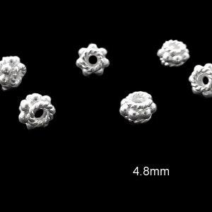 Può includere: Primo piano di diverse piccole perline color argento su sfondo nero. Le perline hanno un design strutturato a forma di fiore con un foro centrale. La dimensione delle perline è di 4,8 mm.