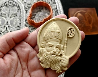 May include: A beige, round, decorative plaque with a raised image of a bearded man wearing a bishop's hat and holding a staff. The man's face is detailed and the staff has a decorative top.