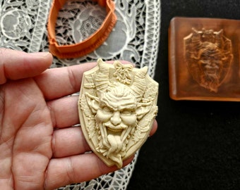 Puede incluir: Un molde de jabón beige en forma de escudo con un relieve detallado de un demonio cornudo y sonriente con la lengua fuera. Un cortador de galletas naranja y una pastilla de jabón marrón con un diseño similar también son visibles.