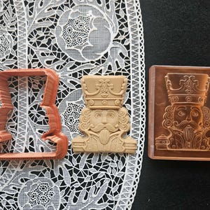May include: Three items related to a nutcracker: a cookie cutter, a cookie, and a stamp. The cookie cutter is a light orange color. The cookie is tan and has a detailed nutcracker face. The stamp is brown and has a detailed nutcracker face.