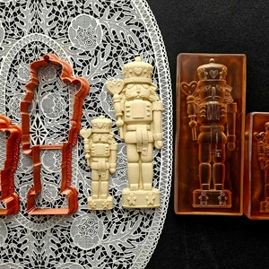 May include: A selection of Christmas-themed items, including two orange plastic cookie cutters shaped like a nutcracker, two off-white nutcracker figurines, and two brown nutcracker moulds.