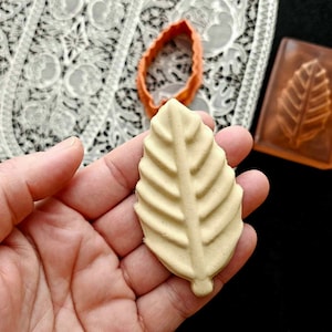 May include: A beige leaf-shaped cookie held in a hand, with a detailed vein pattern. An orange leaf-shaped cookie cutter and a leaf-imprinted mold are also visible. The background includes a white lace pattern and a black surface.