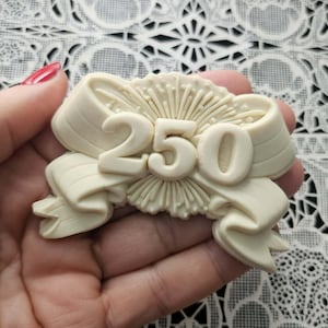 May include: A cream-colored decorative bow with the number "250" in the center. The bow has a ribbon design with detailed folds and a sunburst-like pattern behind the numbers. The object is held in a hand.