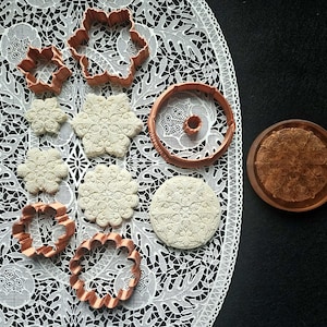 May include: An assortment of copper-colored cookie cutters in various floral and geometric shapes, alongside stamped cookies. The cookies are light-colored with detailed embossed designs. The items are arranged on a white lace tablecloth and a black surface.