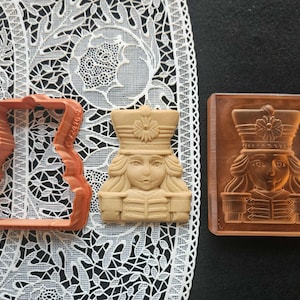 Puede incluir: Un cortador de galletas de plástico naranja y un sello de galletas detallado, ambos con un diseño de soldado. El sello es rectangular y muestra la cara y el uniforme del soldado. El cortador de galletas es el contorno del soldado. Fondo: mantel de encaje blanco y superficie negra.