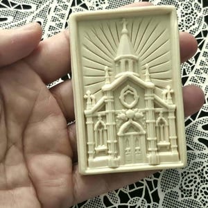 May include: A rectangular, off-white soap bar featuring a detailed, raised relief design of a church building. The architectural depiction includes a central steeple, arched windows, and a doorway, all set against a sunburst pattern background.