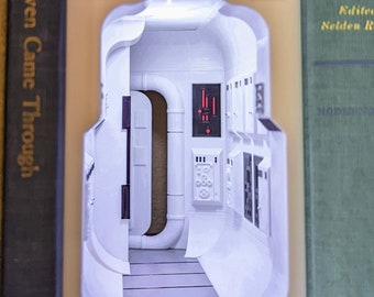 Puede incluir: Un marcapáginas blanco y beige impreso en 3D con forma de interior futurista, con una serie de puertas, paneles y una escalera. El marcapáginas está colocado entre las páginas de un libro con una cubierta oscura.