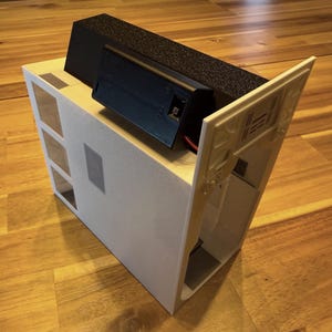 May include: A white rectangular housing unit featuring a series of square cutouts on one side and a black battery compartment mounted on the top. The structure includes an angled white panel attached to the side with visible internal support ribs.