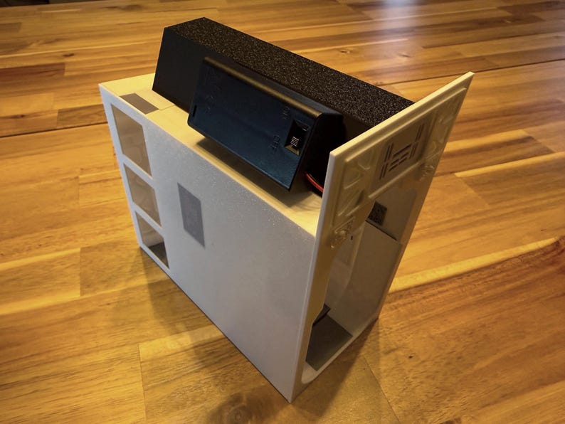 May include: A white rectangular housing unit featuring a series of square cutouts on one side and a black battery compartment mounted on the top. The structure includes an angled white panel attached to the side with visible internal support ribs.