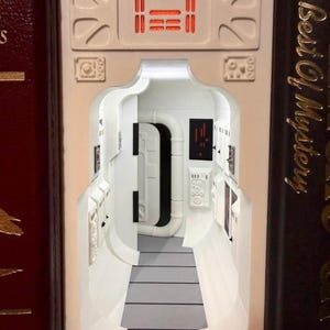 May include: A book nook insert featuring a white sci-fi corridor with a gray floor, a black door frame, and control panels with red and black details. The structure is positioned between two books, one with the text The Best Of Mystery visible.