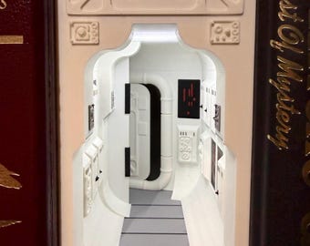 May include: A book nook insert featuring a white sci-fi corridor with a gray floor, a black door frame, and control panels with red and black details. The structure is positioned between two books, one with the text The Best Of Mystery visible.