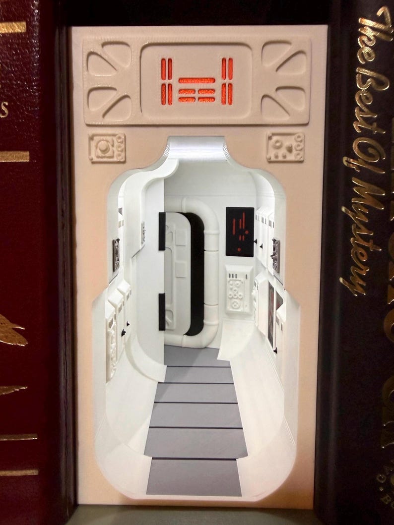 May include: A book nook insert featuring a white sci-fi corridor with a gray floor, a black door frame, and control panels with red and black details. The structure is positioned between two books, one with the text The Best Of Mystery visible.