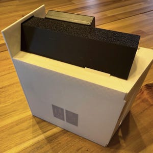 May include: A white rectangular box with an angled top edge, featuring a black rectangular insert with a textured surface. The front of the white box displays two small, light gray square icons positioned side by side in the lower center area.