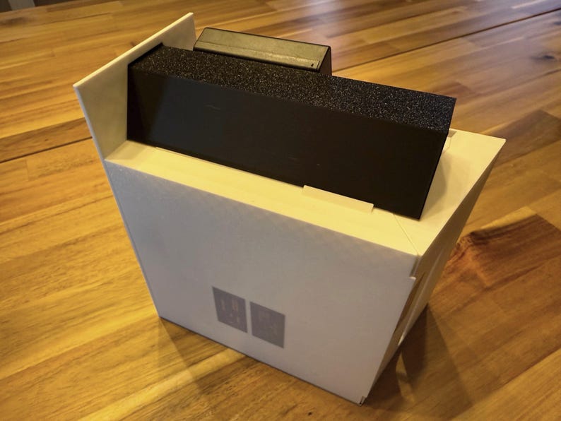 May include: A white rectangular box with an angled top edge, featuring a black rectangular insert with a textured surface. The front of the white box displays two small, light gray square icons positioned side by side in the lower center area.