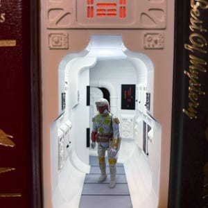 May include: A white 3D-printed book nook insert featuring a sci-fi corridor with illuminated red panels and a detailed figurine of a bounty hunter in green, yellow, and red armor standing in the center of the hallway between two dark books.