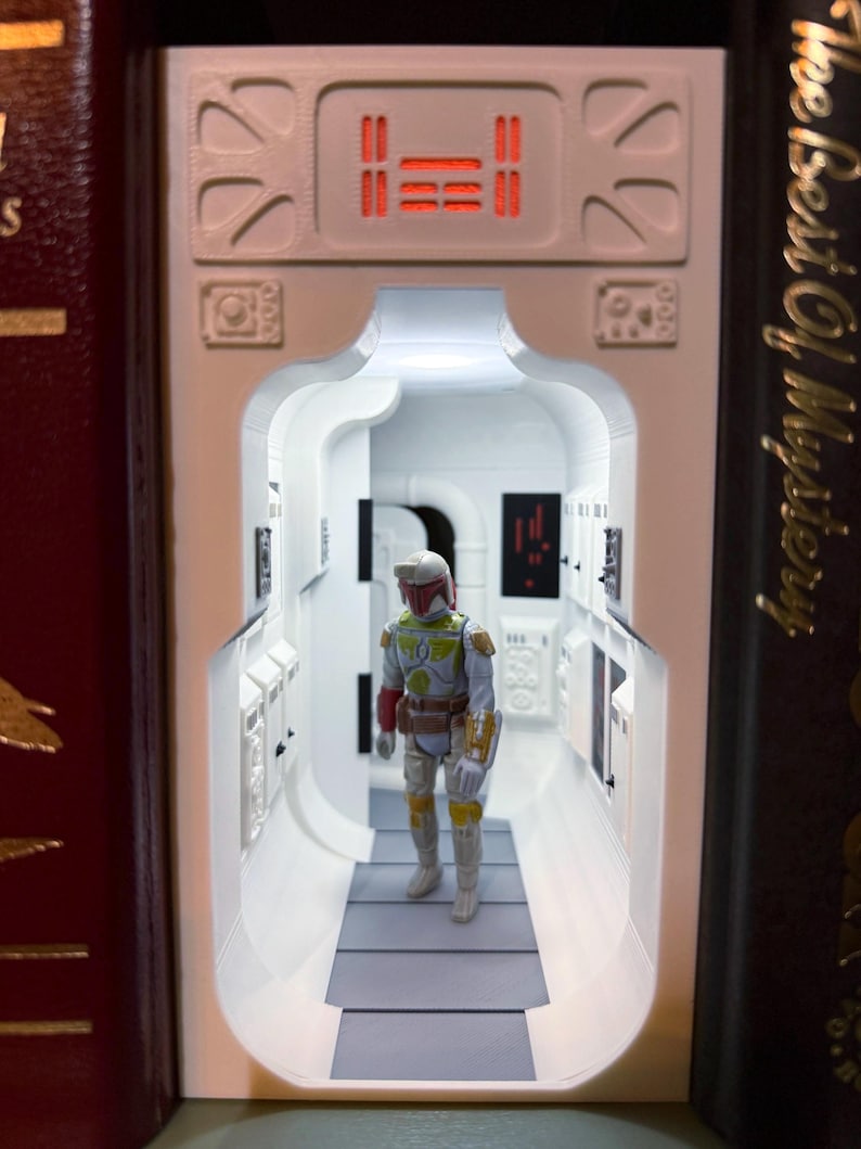 May include: A white 3D-printed book nook insert featuring a sci-fi corridor with illuminated red panels and a detailed figurine of a bounty hunter in green, yellow, and red armor standing in the center of the hallway between two dark books.