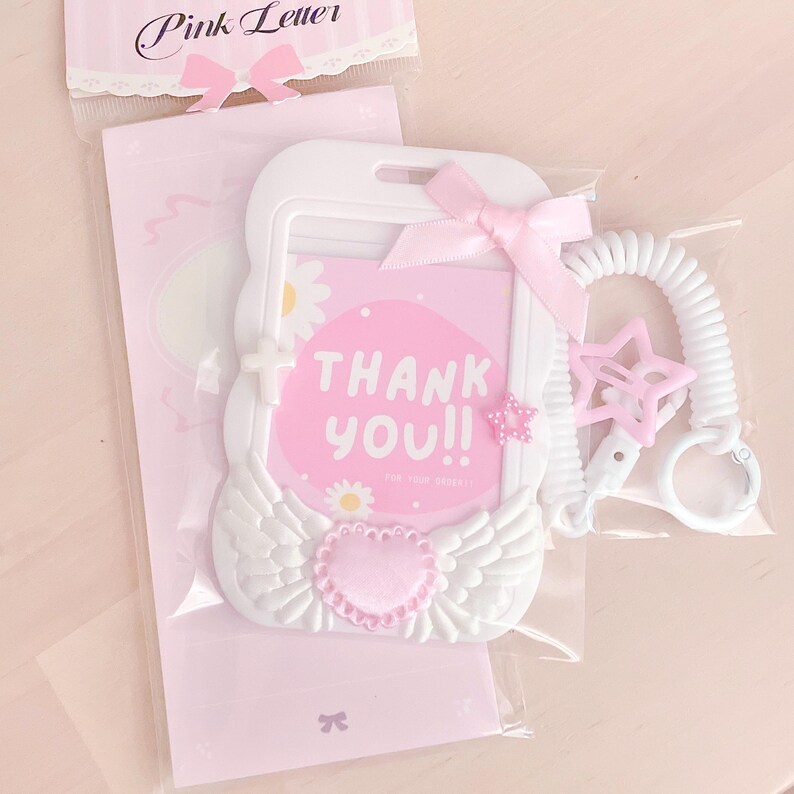May include: White plastic ID card holder with a pink ribbon, a pink heart, and white wings. The holder has a clear window for a card and says "Thank You!" in pink letters. It comes with a white retractable cord and a pink star charm.