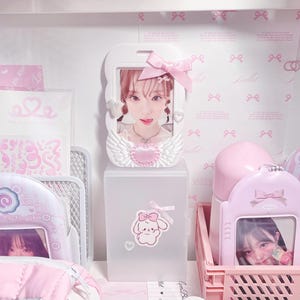 May include: A pink and white plastic storage box with a clear front panel. The box has a small sticker of a cartoon bunny with a pink bow on it. The box is filled with various items, including a small pink and white picture frame with a photo of a young woman, a pink and white water bottle, and a pink and white plastic basket.
