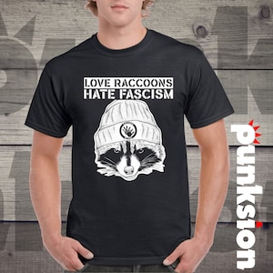 Könnte beinhalten: Schwarzes T-Shirt mit einem weißen Grafikdruck eines Waschbären, der eine Mütze trägt. Der Text "LOVE RACCOONS HATE FASCISM" ist auf dem Shirt aufgedruckt.